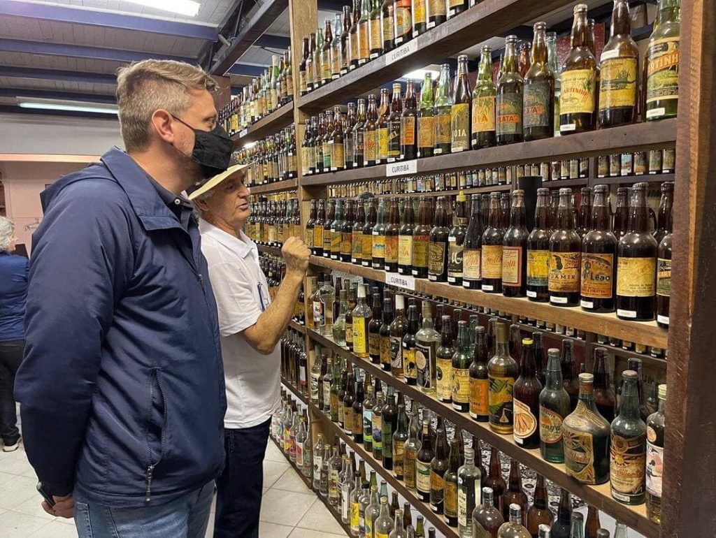 Colecionador de cachaças Orlando mostra acervo para visitantes, no fundo prateleiras de madeira cheias de garrafas de cachaça