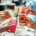 Foto pessoas à mesa fazendo um brinde com copos de tamanhos diferentes e com bebidas diversas e coloridas