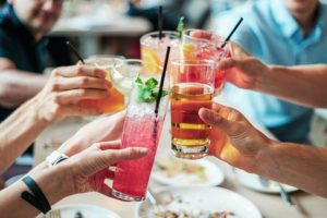 Foto pessoas à mesa fazendo um brinde com copos de tamanhos diferentes e com bebidas diversas e coloridas