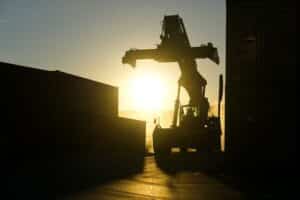Foto contra o sol de um caminhão tipo grua se aproximando de uma pilha de containers